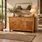 A rustic wooden sideboard or buffet table with two top drawers and two lower cabinet doors, styled in a warm, sunlit room with a lamp, books, a ceramic pitcher, and framed landscape art.