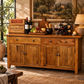 A rustic wooden sideboard or buffet cabinet with two upper drawers and two lower doors, styled in a warm, sunlit dining room. The cabinet is decorated with wine bottles, cheese, grapes, glasses of red wine, a candle, and a wine rack, set against a backdrop of a framed landscape painting and a lamp.