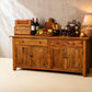 A rustic wooden sideboard or buffet table with four cabinet doors and two drawers, styled with wine bottles, glasses, cheese, grapes, and a candle against a textured beige wall.