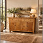 A rustic wooden sideboard or buffet cabinet with four doors and two drawers, styled in a warm, sunlit room with a lamp, books, and potted plants on top.