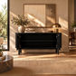 A black wooden sideboard cabinet with open doors, styled in a warm, sunlit room with a gold table lamp, books, and a vase of greenery. A large abstract painting hangs on the beige wall behind it.