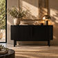 A modern black ribbed sideboard or console cabinet in a warm, sunlit room, styled with a ceramic vase of greenery, a brass dome lamp, and a small bowl.
