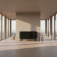 A modern black ribbed sideboard cabinet with slim legs, positioned in a minimalist, sunlit room with high windows and a beige wall behind it.