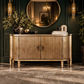 A curved sideboard cabinet with vertical fluted detailing in a warm gold finish, styled in a luxurious room with a dark green paneled wall, a large round mirror, and decorative plants.