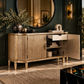 A luxurious gold-finished sideboard cabinet with fluted doors, a single open drawer, and an open side door revealing shelves, styled in an elegant room with a dark green wall, round mirror, and decorative plants.