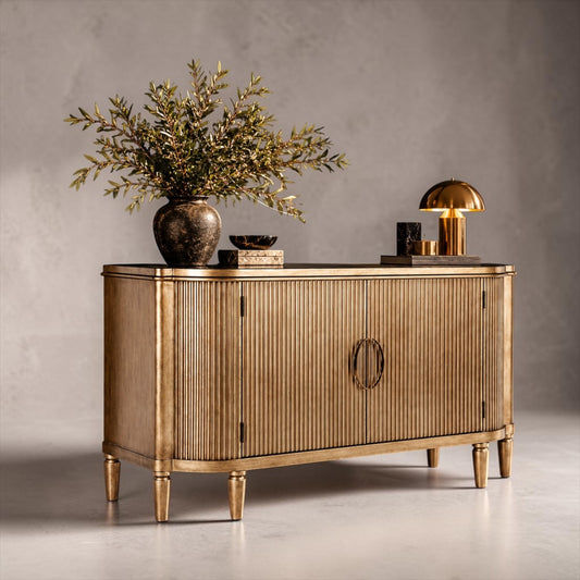 A gold-finished sideboard cabinet with vertical fluting and ring pulls, styled with a vase of greenery and a brass table lamp against a textured gray wall.