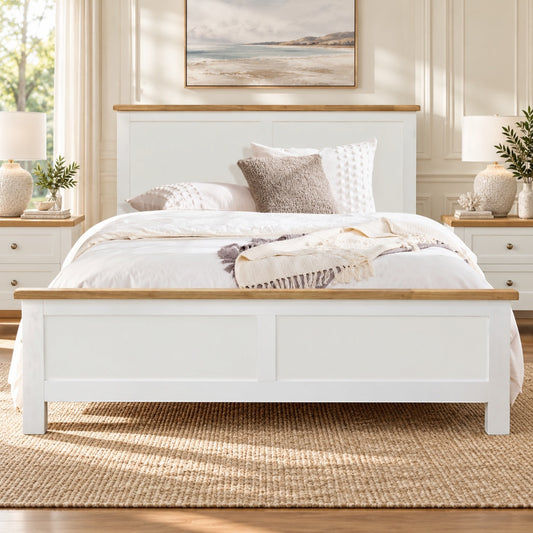 A white wooden bed frame with natural oak tops on the headboard and footboard, styled in a bright, neutral-toned bedroom with white bedding, textured pillows, and a woven jute rug.