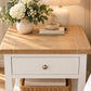 A white nightstand with a light oak wood top, featuring a single drawer with a round metal knob and a woven basket shelf. The nightstand is styled with a ceramic vase of white flowers, a ribbed ceramic table lamp, and a small stack of books in a warm, sunlit room.