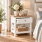A white wooden nightstand with a light oak top, styled in a bright bedroom with a vase of white flowers, a ceramic table lamp, and a woven basket on the shelf.