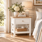 A white nightstand with a light wood top, featuring a single drawer with a round knob and an open lower shelf with crisscross legs. The nightstand is styled in a bedroom setting with a lamp, a vase of white flowers, books, and a woven basket on top and inside the shelf.
