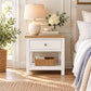A white nightstand with a light wood top, featuring a single drawer with a round brass knob and an open lower shelf holding a woven basket. The nightstand is styled in a bedroom setting next to a bed with white linens, a beige upholstered headboard, a table lamp, and a vase of white flowers.