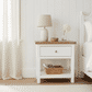A white wooden nightstand with a single drawer and open lower shelf, styled in a bright bedroom with a lamp, vase, and basket. The nightstand is positioned next to a white bed against a white paneled wall.
