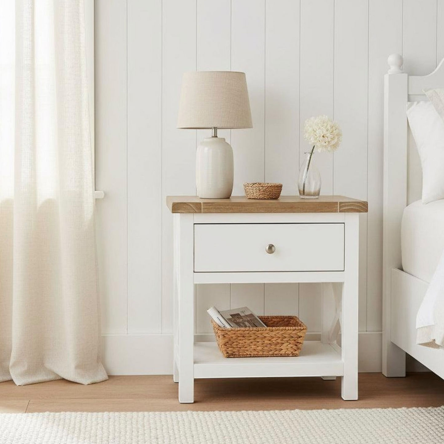 A white wooden nightstand with a single drawer and open lower shelf, styled in a bright bedroom with a lamp, vase, and basket. The nightstand is positioned next to a white bed against a white paneled wall.