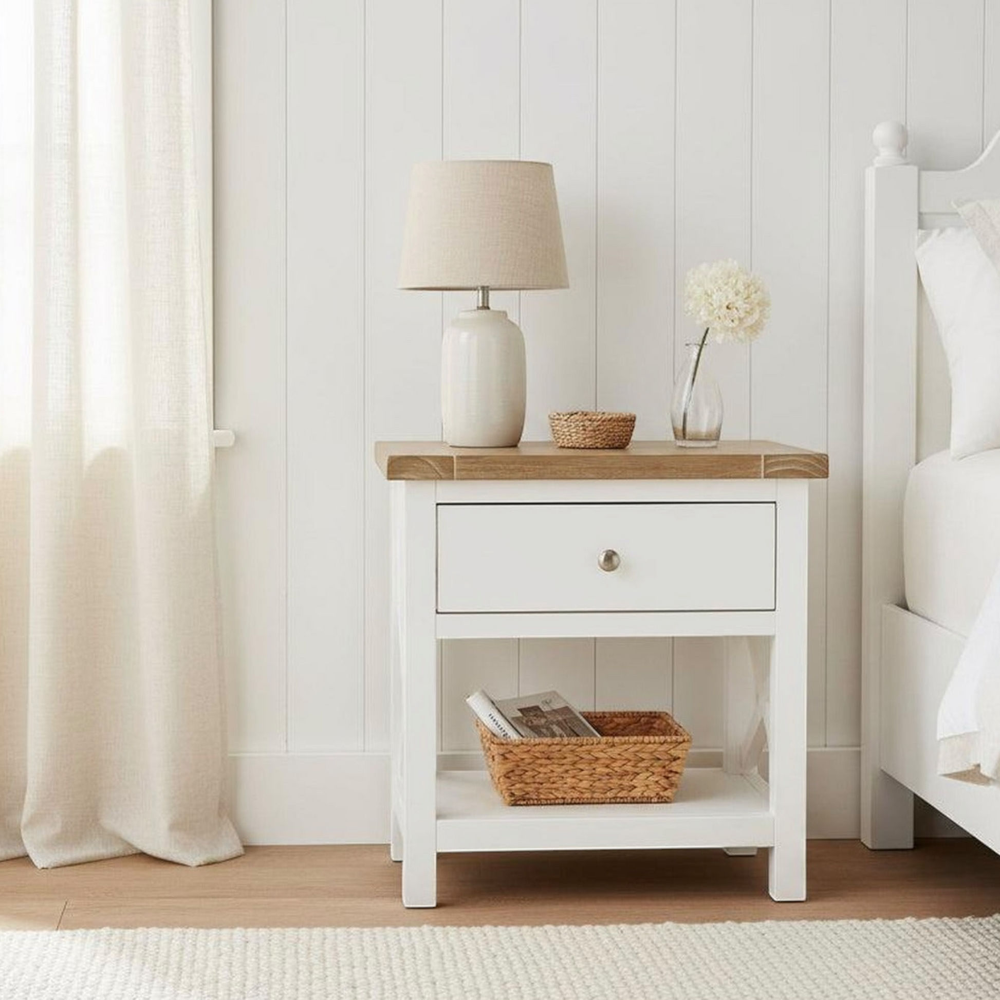 A white wooden nightstand with a single drawer and open lower shelf, styled in a bright bedroom with a lamp, vase, and basket. The nightstand is positioned next to a white bed against a white paneled wall.