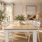 A long dining table with a light natural wood top and white legs, surrounded by white dining chairs in a bright, neutral-toned dining room. A potted olive tree sits on the table, and the room features cream walls, a framed landscape print, and warm natural light from a window.
