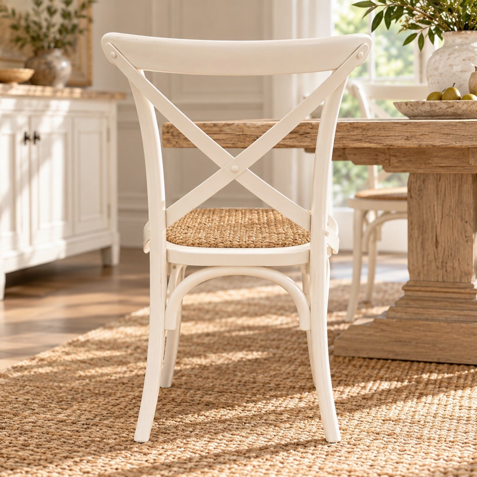 A white wooden dining chair with a woven cane seat, positioned in a bright, sunlit dining room next to a rustic wooden table on a jute rug.