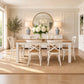 A dining room scene showing a white dining table with six matching white chairs, set on a woven jute rug in a bright, neutral-toned room with a round mirror, lamps, and floral centerpiece.