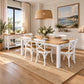 A dining room scene showing a rectangular wooden dining table with six white chairs, set on a large woven jute rug in a bright, coastal-themed room with a window, sideboard, and a framed ocean landscape painting on the wall.