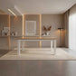A modern rectangular dining table with a light wood top and white legs, set on a cream rug in a minimalist, warm-toned dining room.