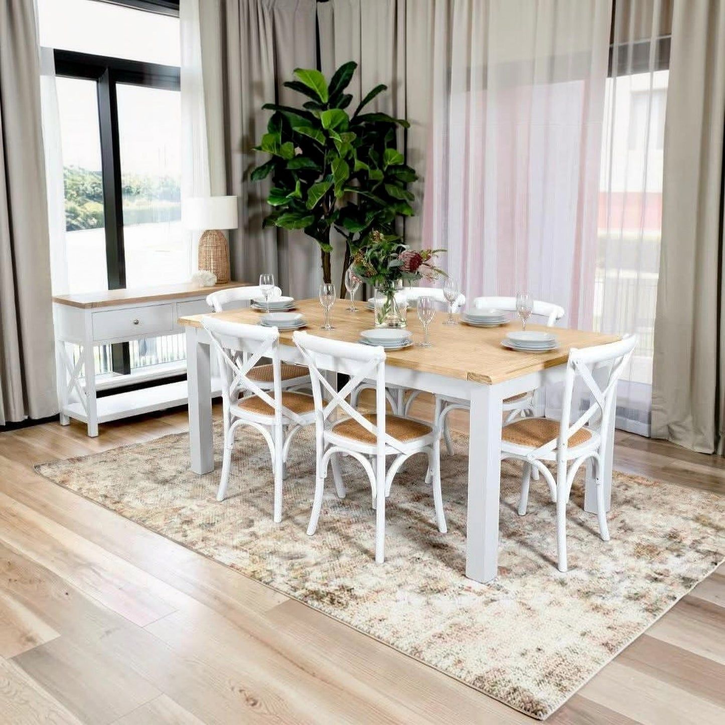 A dining room scene showing a rectangular table with a light natural wood top and white legs, surrounded by six white chairs with wooden seats. The table and chairs are placed on a large beige patterned area rug in a bright room with wood floors, sheer curtains, and a potted plant in the background.