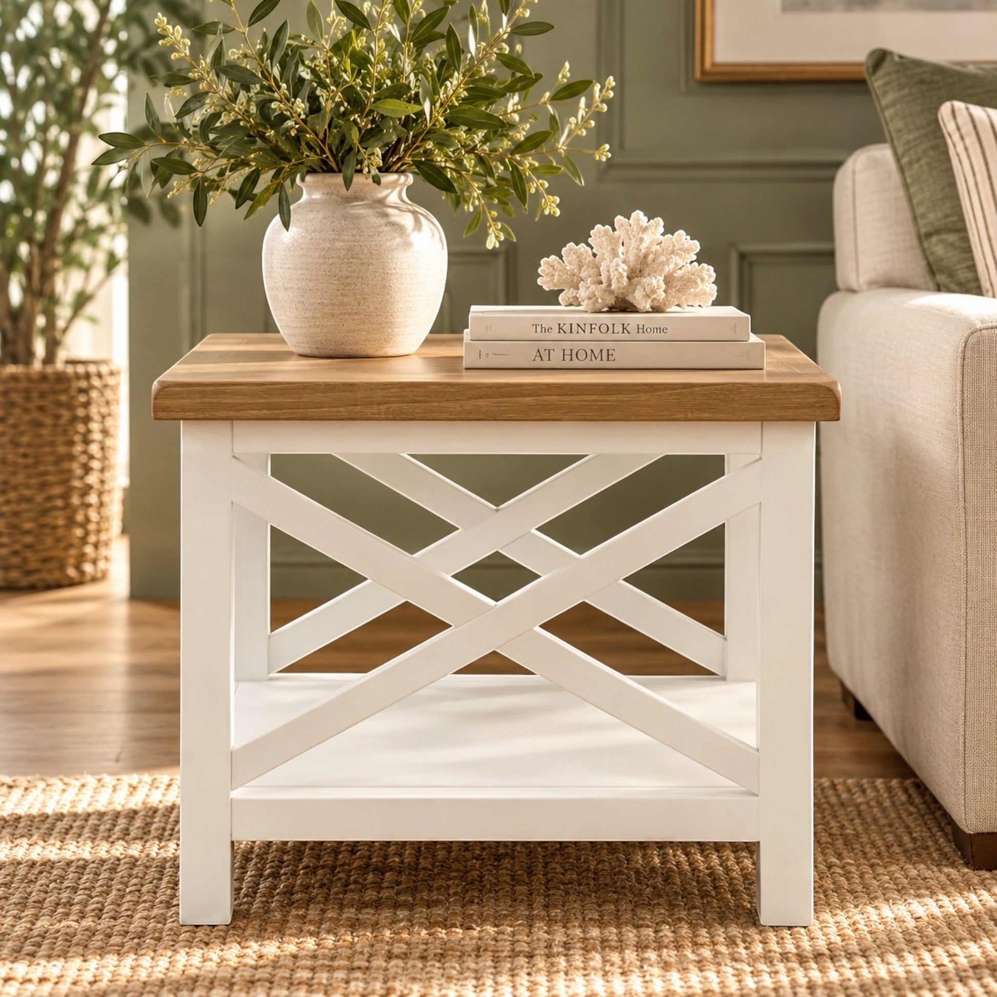 A white wooden end table with a natural oak top and an X-shaped lower support frame, styled in a living room with a vase of greenery, books, and a coral sculpture on top.