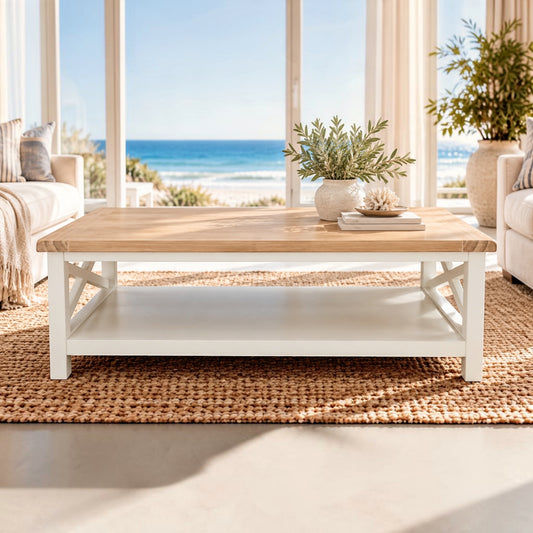 A rectangular coffee table with a light natural wood top and a white frame, featuring an open lower shelf. The table is styled on a woven jute rug in a bright, modern living room with large windows overlooking the ocean.