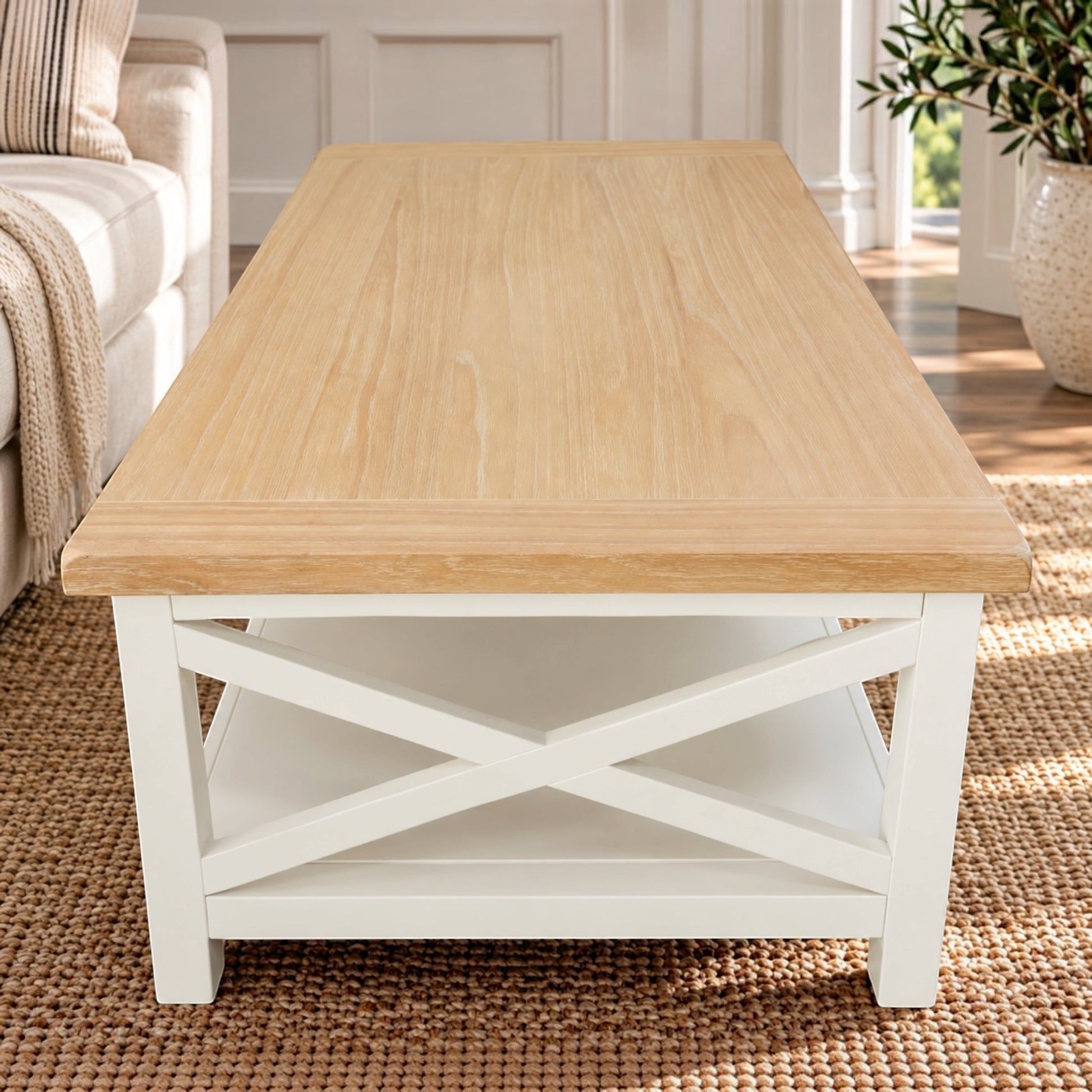 A rectangular coffee table with a light natural wood top and a white frame featuring an X-shaped lower shelf, shown in a bright living room setting.