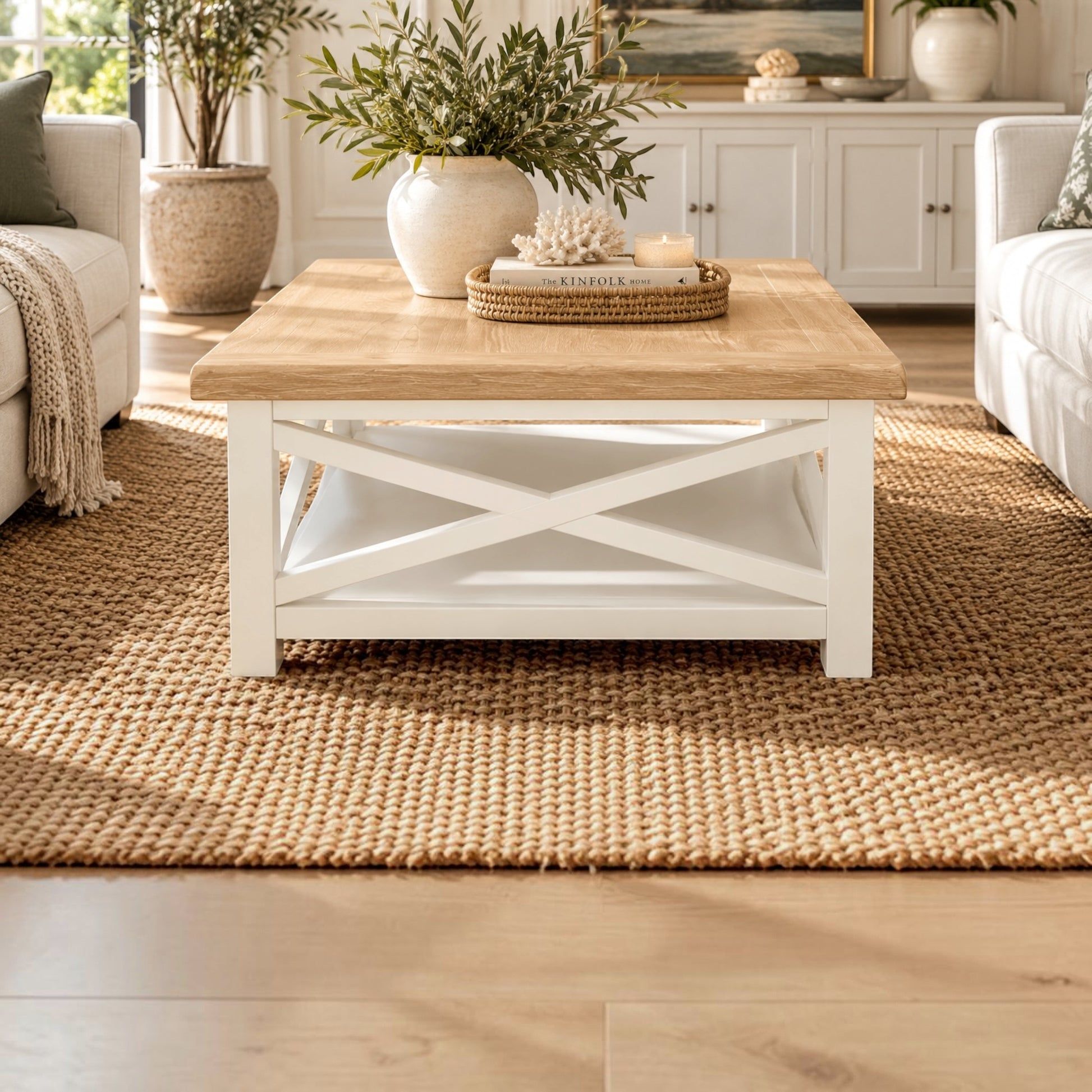 A white wooden coffee table with a light natural wood top and an X-shaped lower shelf, styled in a bright, modern living room on a woven jute rug.