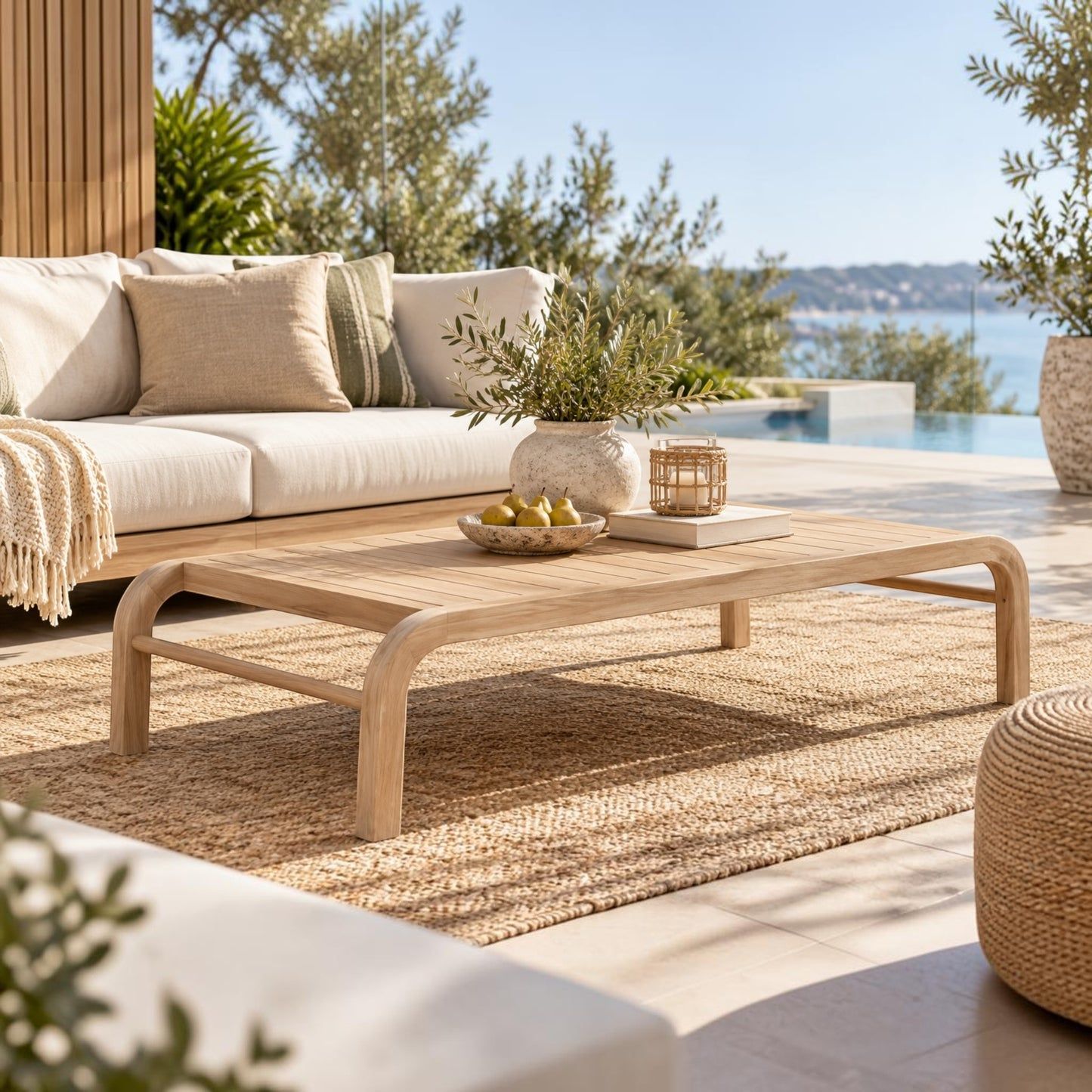 A light natural wood coffee table with curved legs in an outdoor patio setting, styled with a woven rug, a cream sofa, and a view of the sea in the background.