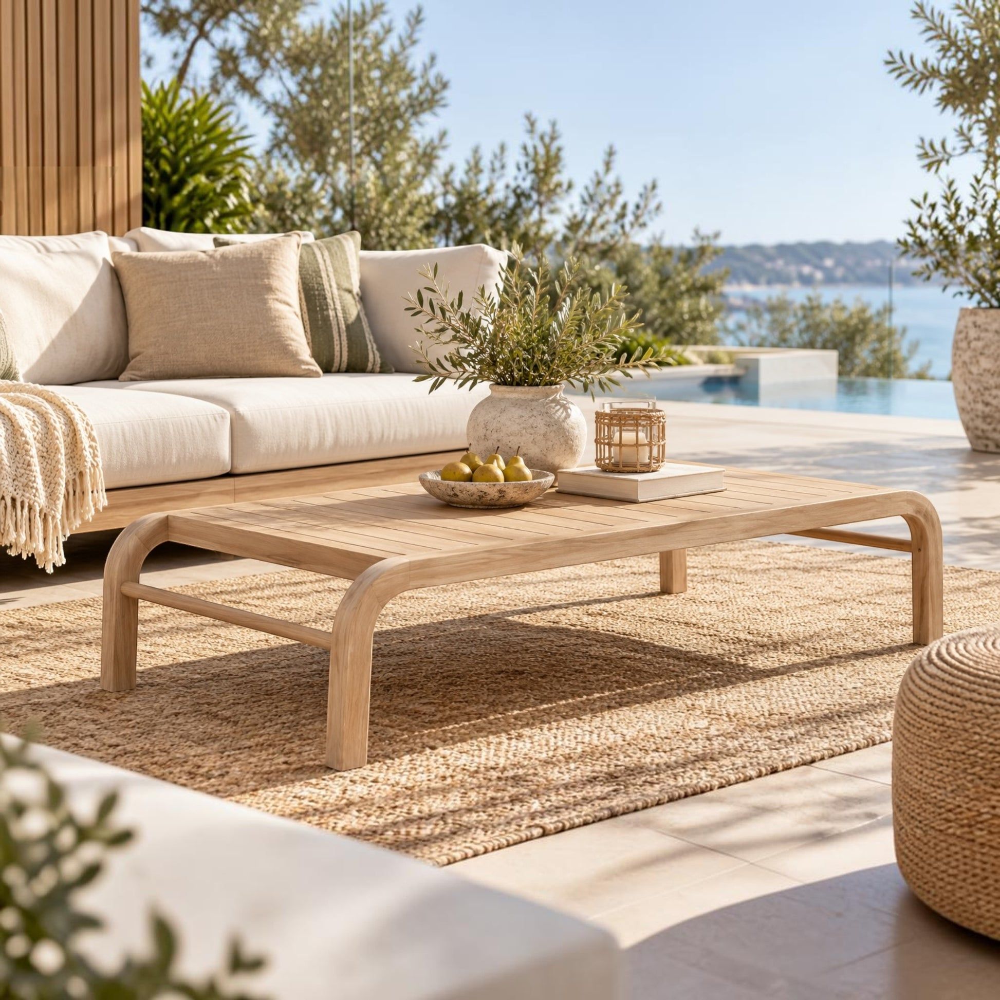A light natural wood coffee table with curved legs in an outdoor patio setting, styled with a woven rug, a cream sofa, and a view of the sea in the background.