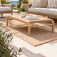 A light natural wood outdoor coffee table with curved legs sits on a woven jute rug in a sunlit patio setting. The table is styled with a small potted plant and a book, with a matching sofa and outdoor seating visible in the background.
