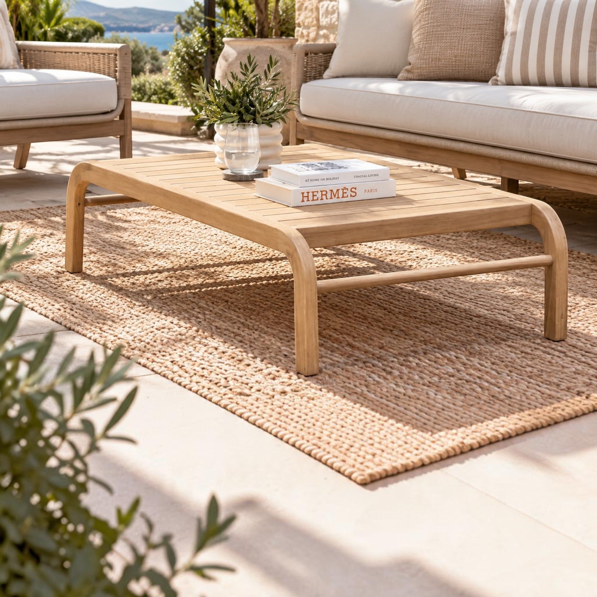 A light natural wood outdoor coffee table with curved legs sits on a woven jute rug in a sunlit patio setting. The table is styled with a small potted plant and a book, with a matching sofa and outdoor seating visible in the background.