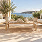 A light natural wood outdoor coffee table with curved legs sits on a woven jute rug on a stone patio, with a blue sea and distant island visible in the background. Matching outdoor furniture and potted plants surround it.