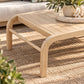 A light natural wood outdoor coffee table with curved legs and a slatted top, styled on a woven beige rug with a potted plant on a tray in the corner.