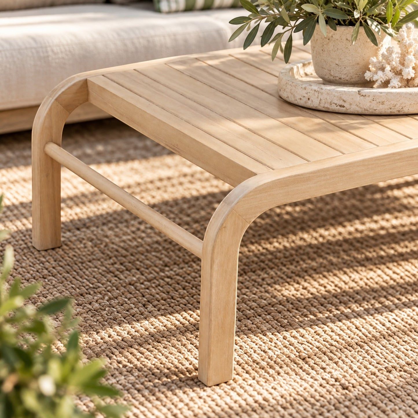 A light natural wood outdoor coffee table with curved legs and a slatted top, styled on a woven beige rug with a potted plant on a tray in the corner.