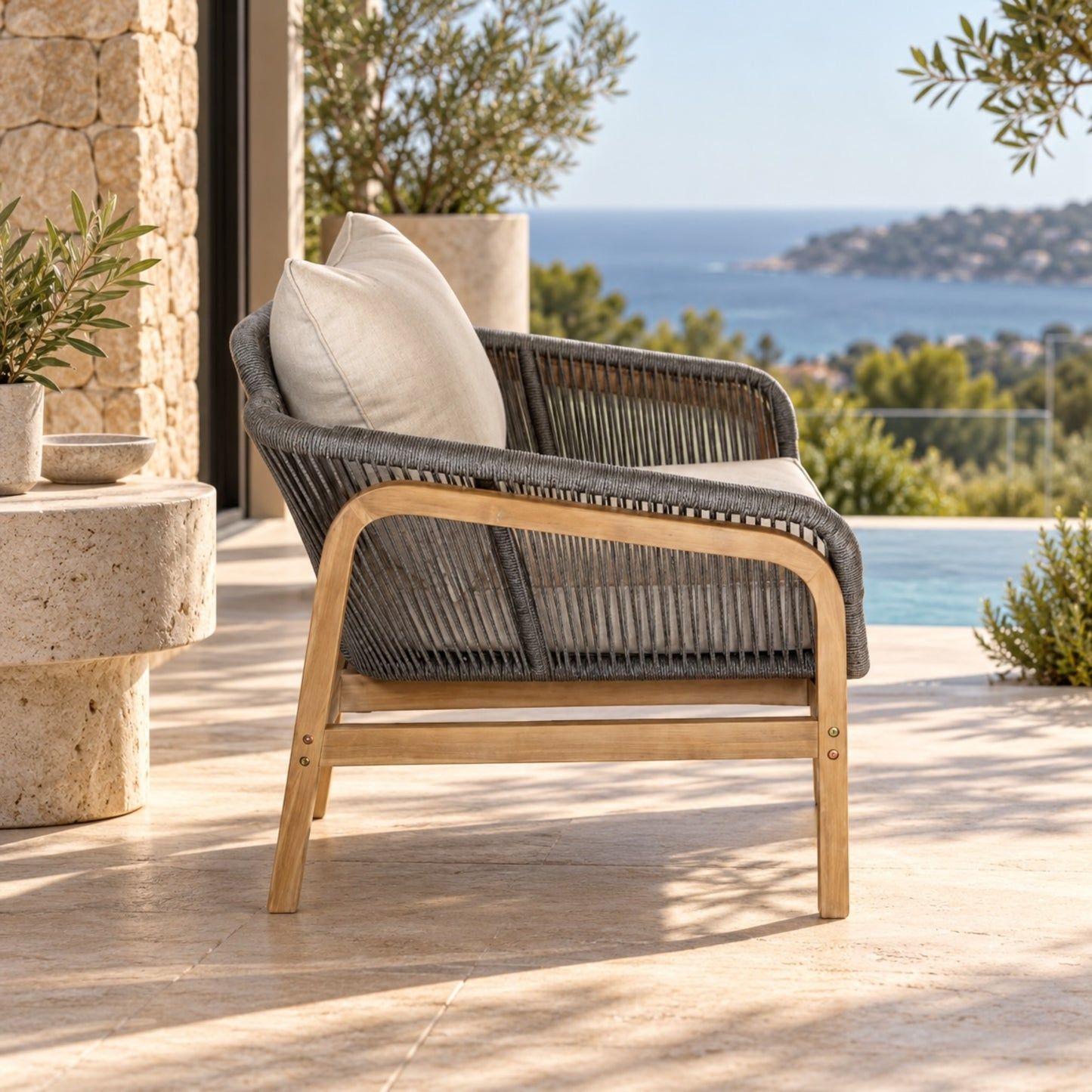 A modern outdoor lounge chair with a gray woven rope seat and back, light natural wood frame, and off-white cushions, positioned on a stone patio overlooking a pool and sea view.