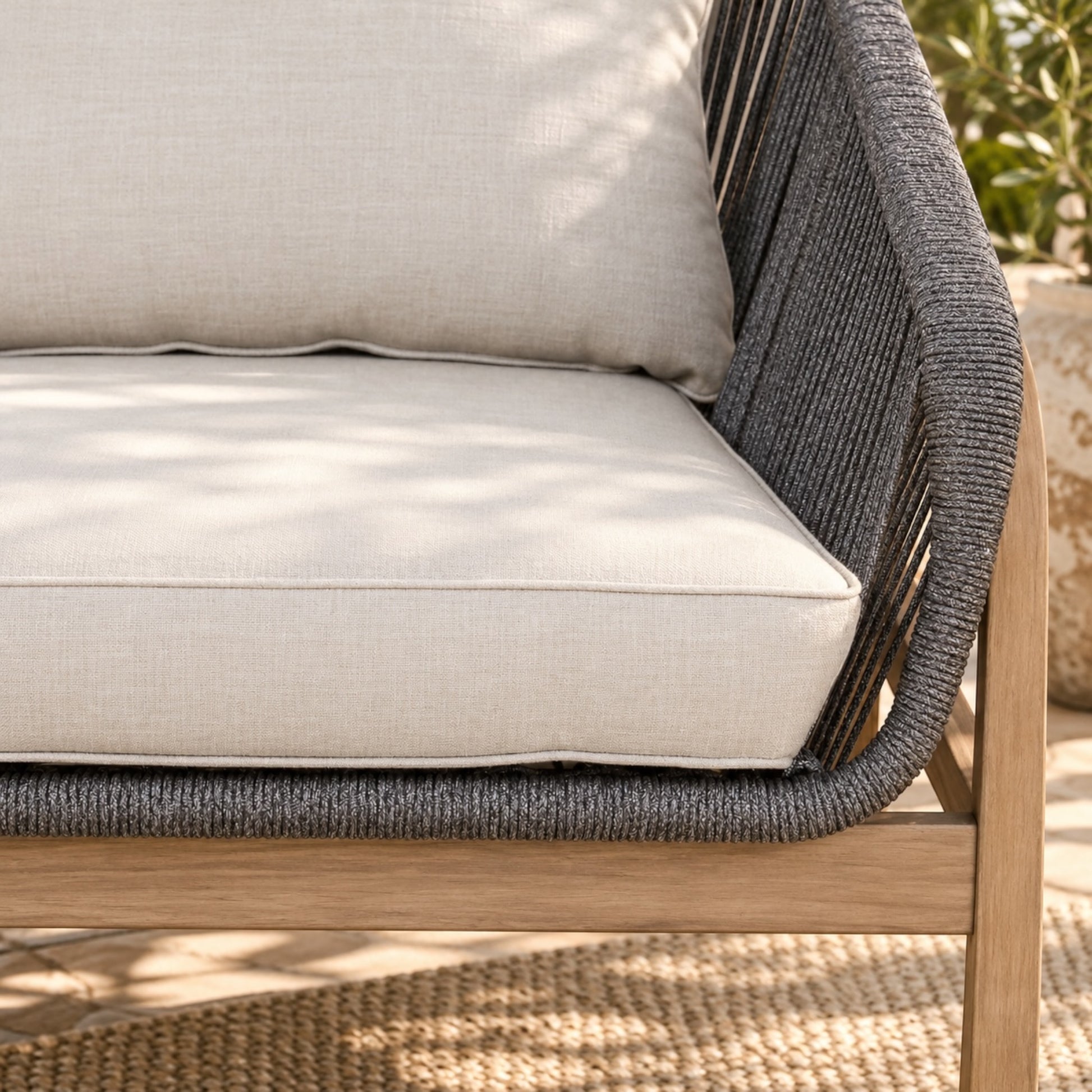 Close-up of an outdoor sofa cushion set with a light beige fabric seat cushion, back cushion, and dark gray woven rope armrests on a light wood frame.