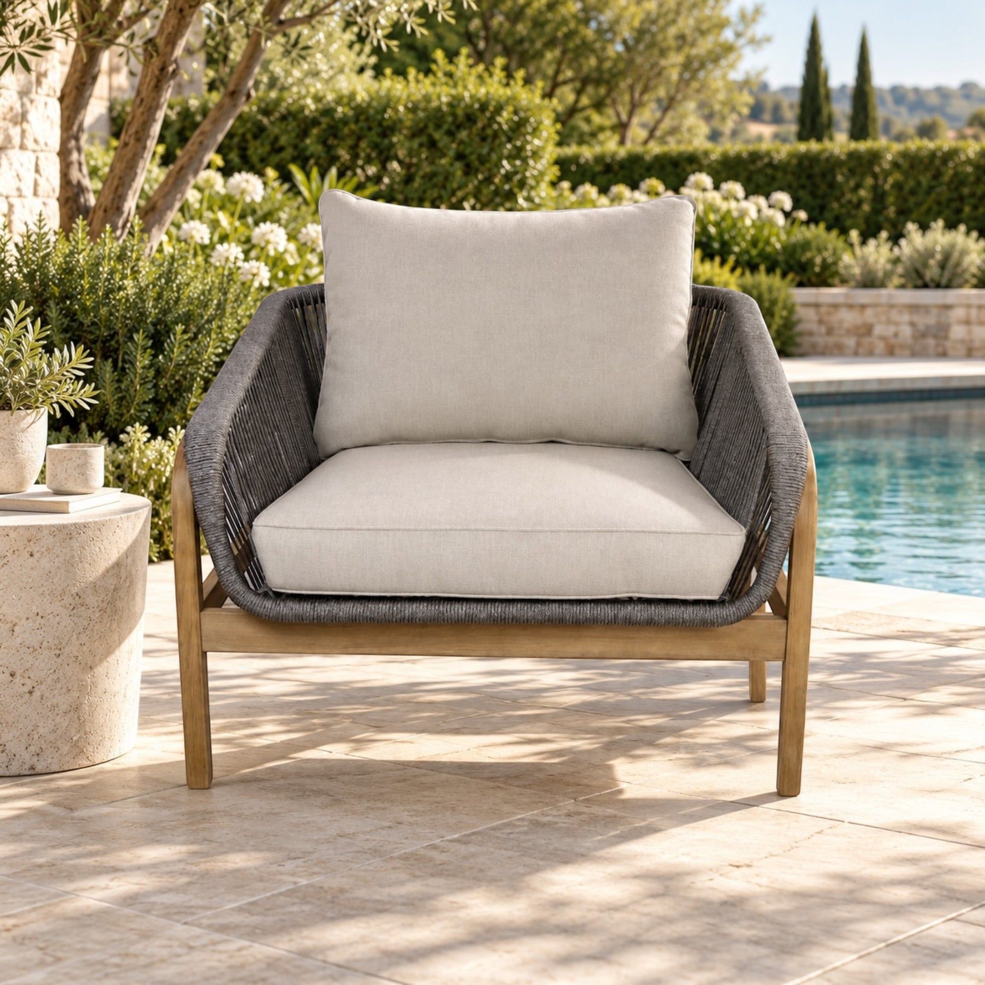 Outdoor patio armchair with a light beige cushioned seat and backrest, dark gray woven rope sides, and a light wood frame, positioned beside a swimming pool on a stone patio.
