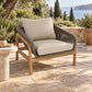 Outdoor patio armchair with a light wood frame, woven gray rope arms, and thick beige cushions, set on a stone patio with a scenic sea and trees in the background.