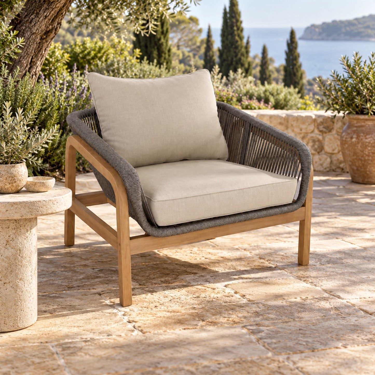 Outdoor patio armchair with a light wood frame, woven gray rope arms, and thick beige cushions, set on a stone patio with a scenic sea and trees in the background.