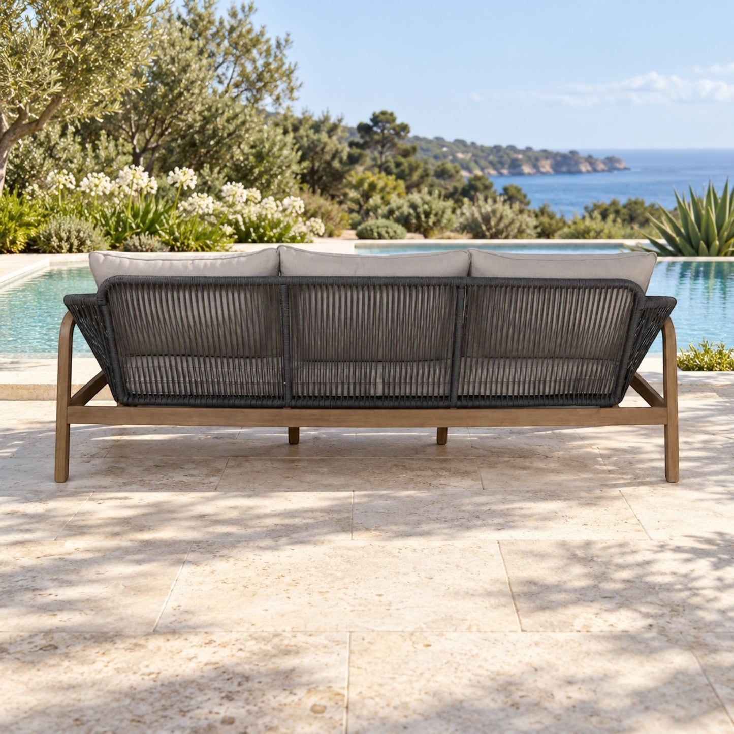 A three-seat outdoor sofa with a dark woven rope backrest and light wood frame, set on a stone patio with a swimming pool and ocean landscape in the background.
