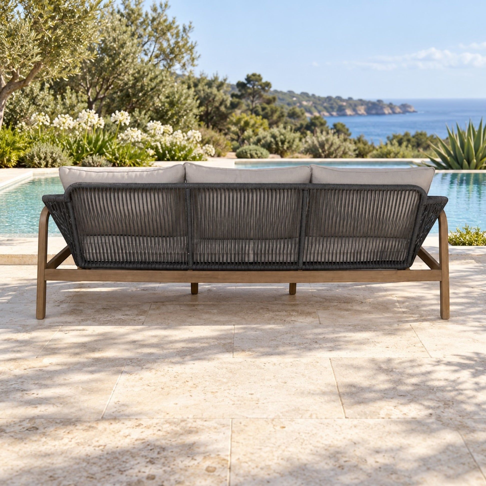 A three-seat outdoor sofa with a dark woven rope backrest and light wood frame, set on a stone patio with a swimming pool and ocean landscape in the background.