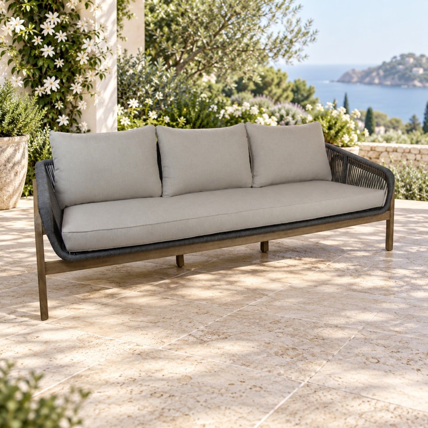 A three-seat outdoor sofa with light beige cushions and a dark gray woven frame, set on a stone patio with a Mediterranean sea and garden background.