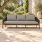 A three-seat outdoor sofa with light beige cushions and a dark woven frame, set on a stone patio with greenery and a building in the background.