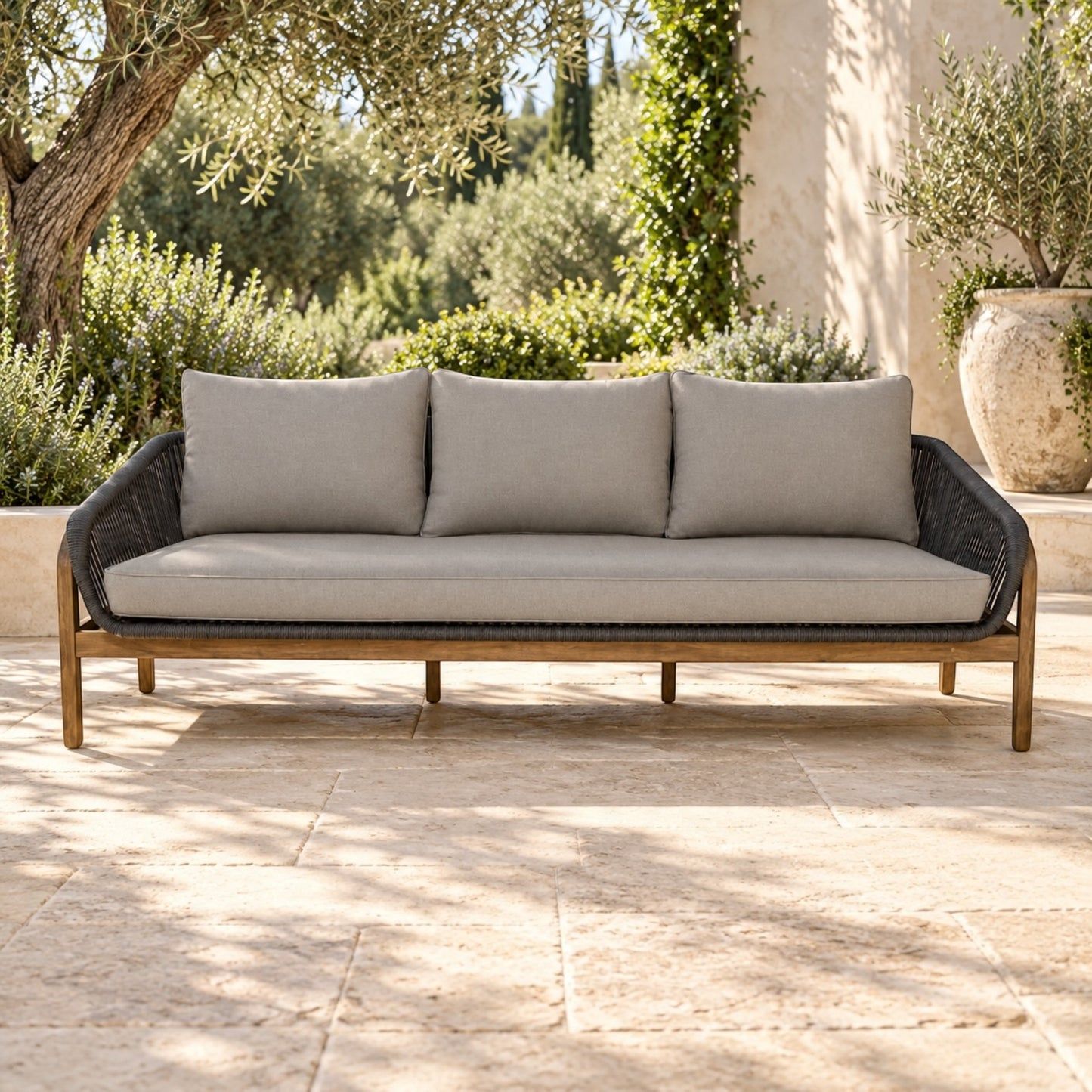 A three-seat outdoor sofa with light beige cushions and a dark woven frame, set on a stone patio with greenery and a building in the background.