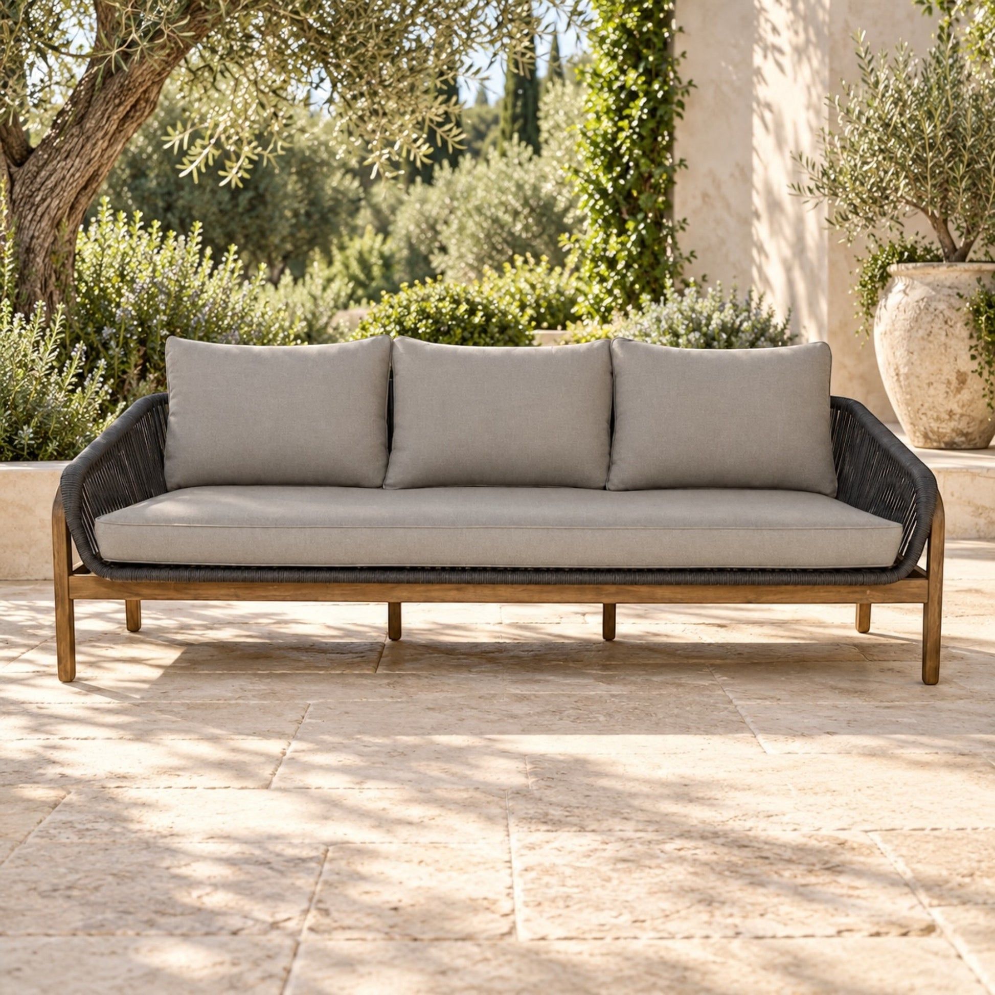 A three-seat outdoor sofa with light beige cushions and a dark woven frame, set on a stone patio with greenery and a building in the background.
