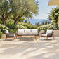 A 4-piece outdoor patio furniture set including a three-seater sofa, two armchairs, and a slatted coffee table, arranged on a stone patio with olive trees, white flowers, and a sea view in the background.