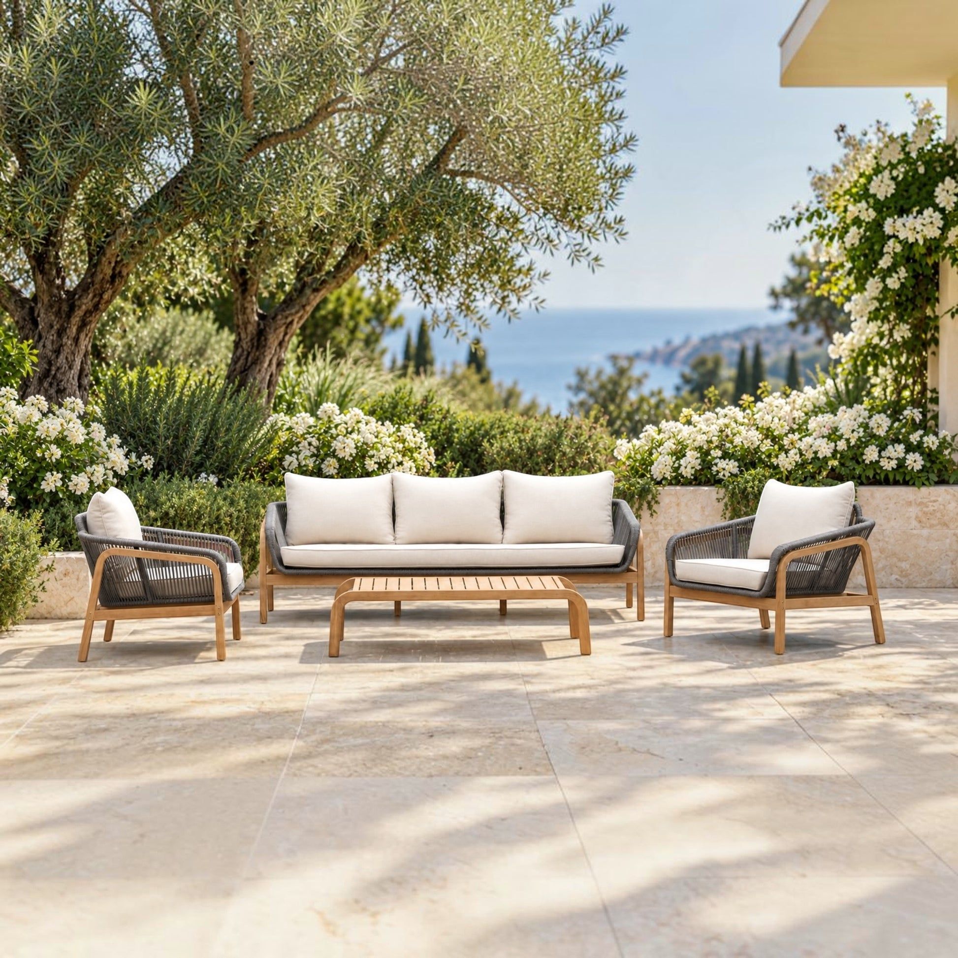 A 4-piece outdoor patio furniture set including a three-seater sofa, two armchairs, and a slatted coffee table, arranged on a stone patio with olive trees, white flowers, and a sea view in the background.