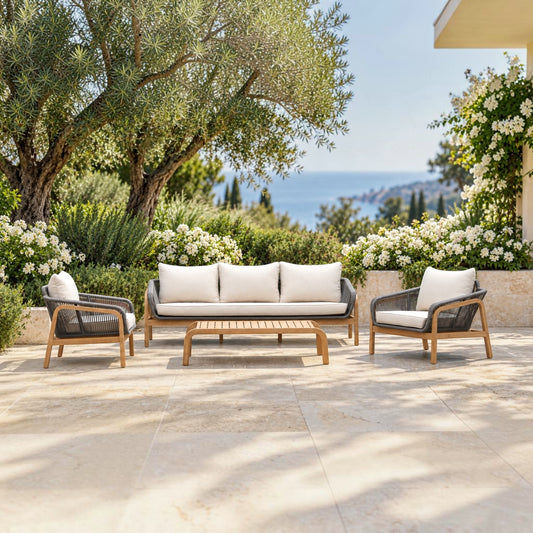 A 4-piece outdoor patio furniture set including a three-seater sofa, two armchairs, and a slatted coffee table, arranged on a stone patio with olive trees, white flowers, and a sea view in the background.
