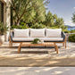 A modern outdoor patio sofa with white cushions and a light wood frame, paired with a matching wooden coffee table on a tiled patio with a woven rug, olive trees, and a scenic ocean view in the background.
